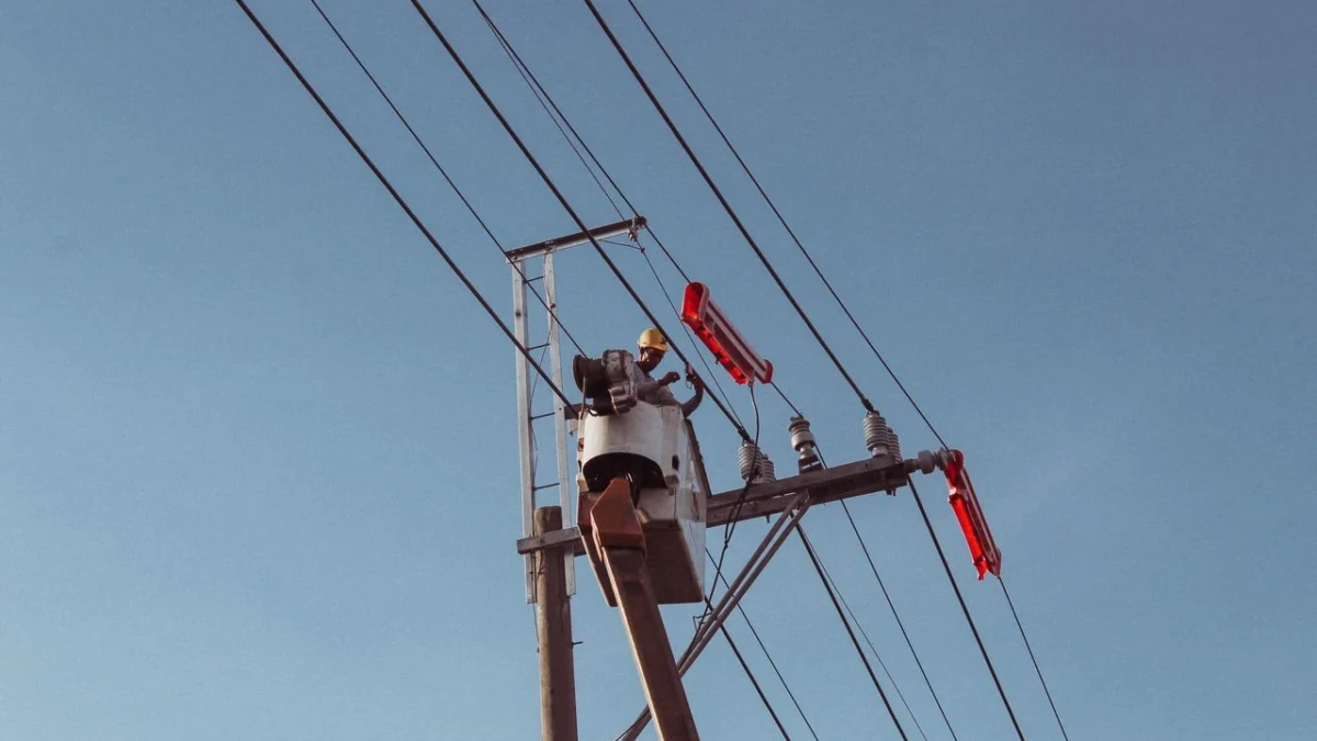 Alçak açıdan enerji direği, sepetli araç platformunda hat üzerinde çalışan baretli tekniker ve gökyüzünü çapraz kesen iletkenler
